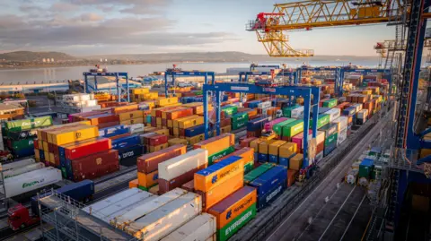 Shipping containers pictured beside the port. The containers are multi-coloured and there are yellow and blue cranes beside them. The lough is in the background. 