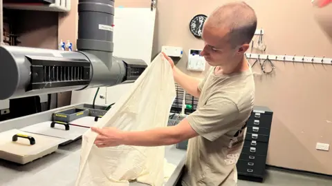 A white male in his twenties, with a shaved head wearing a t-shirt. holding a large latex balloon above a work station in a dark room