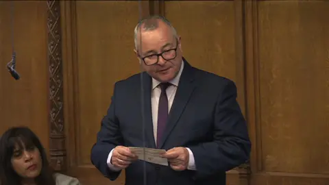 A bespectacled man in a navy suit, dark red tie and white shirt stands and reads off a piece of paper.