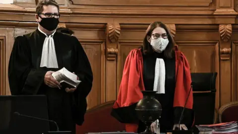 Getty Images Judge Isabelle Fachaux arrives for the trial of French retired surgeon Joel Le Scouarnec in 2020. She has dark hair and glasses and is dressed in red robes. Another officer of the court, a man taller than the judge, stands beside her. He is dressed in black robes. They both wear face masks to protect them against the spread of the Covid-19 virus. 