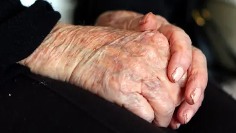 An elderly person's hands that rest in their lap. They wear pink nail varnish which has slightly chipped.
