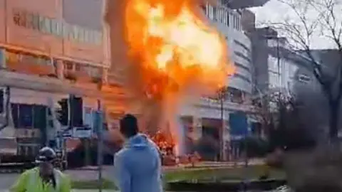 Fireball erupts from truck outside shopping centre as man watches from a distance and another man in high vis runs away