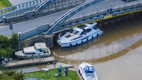 Oliv3r Drone Photography A white cruiser is wedged sideways on underneath a metal bridge.