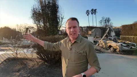 John Sudworth standing in front of a burnt out shell of a car and a house burnt to the ground