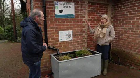 Two people, a man and a woman, stand by a grey metal planter. It is under a drain pipe and contains some plants.