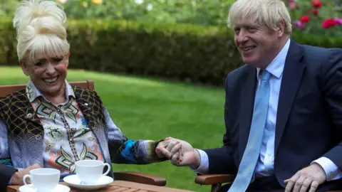 PA Media A woman with blonde hair piled high on her head sits across from a man with blond hair and a blue shirt, tie and suit. They have two white tea cups in front of them 