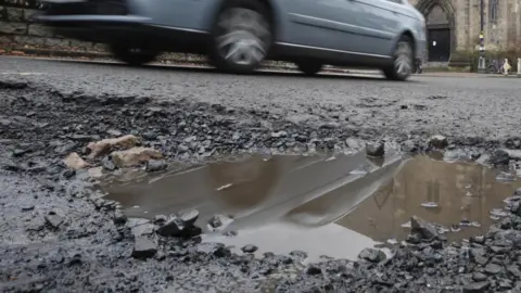 A ground-level image of a pothole in a road, full of water. A light blue car is driving past in the background. The entrance to a building is also visible in the distance.