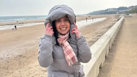 Kate Dawson is standing on a beach promenade with a big beach and the sea behind her. She is wearing a grey coat and a pink scarf 