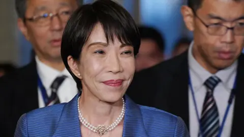 Japan's Prime Minister Sanae Takaichi, in a pearl necklace and blue striped blazer, smiles as she arrives for a debate with opposition party leaders in the parliament building on November 26, 2025