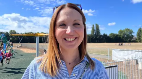 A woman with long brown hair smiles at the camera - she is wearing a blue and white striped blouse and the chain of a handbag can be seen over one shoulder. A playpark is in the background.