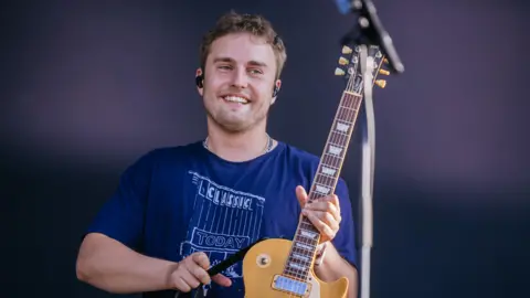 Sam Fender Belfast: Thousands heading to Belfast Vital gig 1 Thomas Niedermueller/Getty Images Sam Fender performing onstage during Southside Festival at Take-off Gewerbepark on 22 June, 2025 in Neuhausen, Germany. He has short, curly brown hair and stubble on his face. He is smiling and holding up a yellow electric guitar. He is weaing a navy t-shirt with a "Classic Today" logo and has a set of black earbuds in his ears.