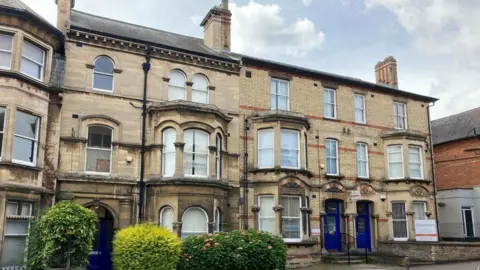 A two-storey block of offices with three blue front doors and bay windows in front of a street pavement and small hedges.