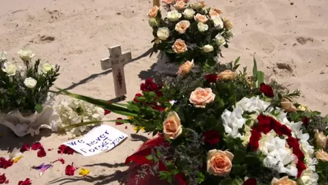 BBC Flowers, tributes and a cardboard cross lie on the sand. A handwritten note on a piece of paper reads: "We will never forget you".