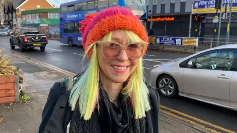 BBC Victoria Platt pictured smiling weating a pink and orange hat and a pair of brown glasses. She has yellow,green and pink hair that is shoulder length and wears a black jacket.