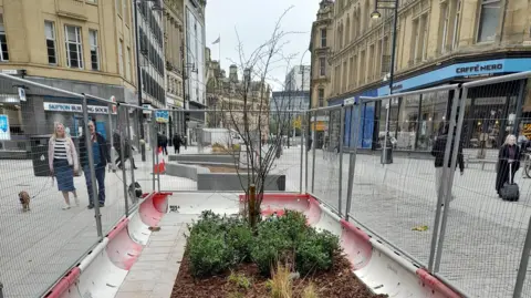 Bradford Council A fenced-off area with a red and white bollard base and green plants surrounding a small tree in a brown soil bed. The area is surrounded by stone buildings.