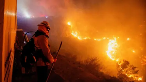 A firefighter stands in front of a large blaze