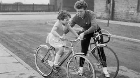 Getty Images A black-and-white photo shows a young child riding a small bicycle on a quiet street. A woman walks alongside, holding the handlebars and guiding the child.