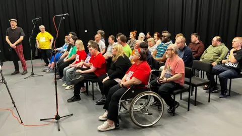 A group of people all sitting in a room on chairs with microphones facing them.