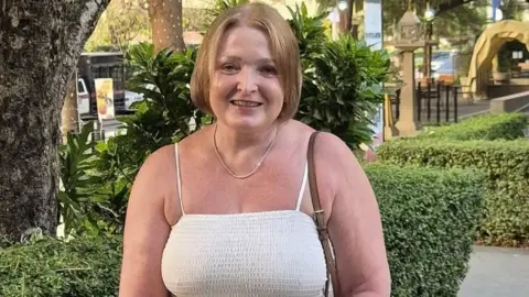 Julie Osgood, pictured in her late 50s, wearing a sleeveless white dress and with light blonde hair cut into a bob, stands in front of a green hedge and smiles at the camera.