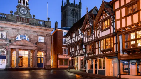 Getty Images A street in the evening with black and white timber buildings along one side of the road and a stone building with arches on the ground floor at one end