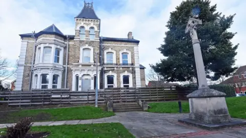 Middlesbrough Council The two-storey villa with a turret has white, arched windows. In front there is a wooden fence with steps leading down a stone monument. 