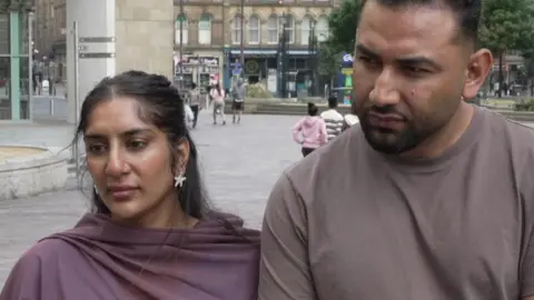 Aisha Iqbal/BBC A young man and woman. She wears a lilac shawl and star shaped earrings. He wears a dark tan t-shirt. They are standing in a paved area with trees behind.