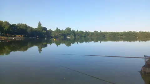 A large flat lake with trees surrounding its far shore and a fishing rod visible on the right hand side