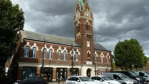 Jonathan Thacker/Geograph March Town Hall is a two storey building which has a bell tower off centre to the right. The bottom floor has different businesses in and there is an entrance underneath the bell tower. There are cars parked outside of the building.