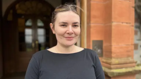 BBC Woman in a black top, and with her hair tied back, standing in front of a sandstone building. 