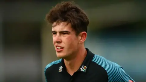 Getty Images Late cricketer Josh Baker, a young man with dark curly hair, looks at something off-camera. He is wearing a black and blue cricketing top.