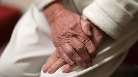 An elderly person rests their hands over one another on their lap. They wear white trousers with a white long sleeved jumper.