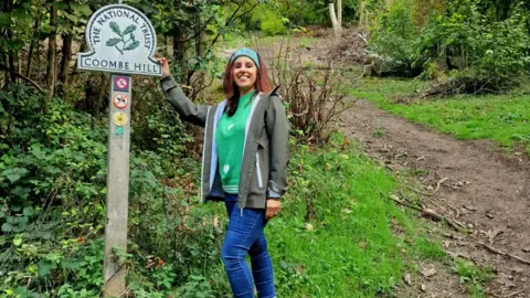 Supplied Geeta standing next to a National Trust sign with its logo and the words "Coombe Hill". It is in woodland and she is holding onto the right side of the sign. She wears blue jeans, a bright green jumper, a sage gren jacket and a turquoise patterned headband. There is a dirt track behind her.