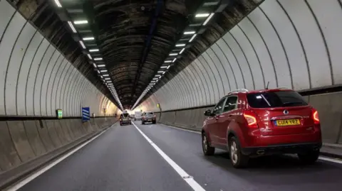 Huw Fairclough/Getty Images Cars driving through a dual carriageway tunnel, with lighting overhead and a long white line through the middle.