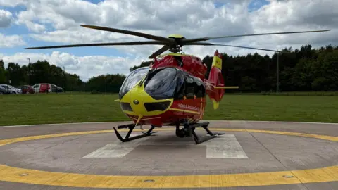 A red and yellow helicopter on a landing pad marked with yellow and white. There is grass and trees behind.