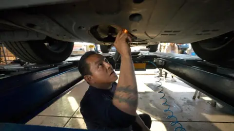 Jorge Gonzalez Jr uses a flashlight to look under a vehicle in an automotive repair shop in Arizona.