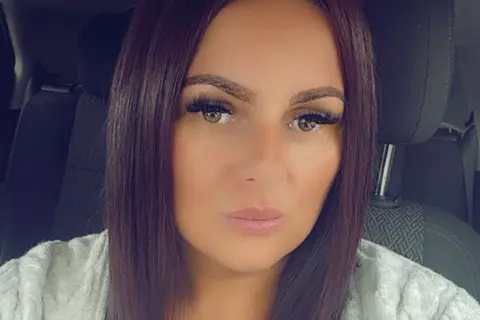 Stacey Lamb Stacey Lamb mira a la cámara una selfie en su auto. Ella tiene cabello longitud de los hombros del color marrón oscuro y lleva un cárdigan gris claro. 