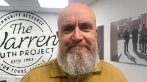 A man in his 50s with a grey and ginger beard smiles with closed lips as he stands in a communal area at a youth cenre. He is wearing a mustard yellow top. Behind him is a white wall with a large, round painted logo that reads: The Warren Youth Project, ESTD 1983, Community Resource for Young People. A canvas with a photograph of young people with skateboards is on a wall to the right. 