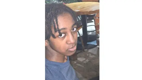 Metropolitan Police A young man with braided medium length dark hair and brown eyes looks into the camera. He is wearing a dark blue t-shirt. There are tables and chairs visible in the background.
