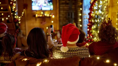 Rear view of multi-generation family watching a movie on TV in a living room surrounded by Christmas decorations
