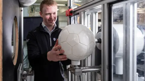 A man wearing a black zip-up jumper holds a white football on a contraption in a lab. He has blonde hair and is smiling slightly. 