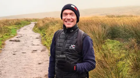 Jay Curtis Jay smiling looking at the camera. He is standing on a muddy walking path on a mountain. Jay wears a long sleeve navy top, a navy hat, a black gilet, black gloves and holds a takeaway coffee in his left hand.