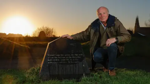 PA Media Michael Burton, an older man with some grey hair and a jacket is crouched at the Stone Circle on the Hill of Infinity as the sun sets close to the winter solstice marker.