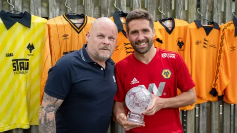 Ed Bagnall One man in a blue polo shirt, one man in a red football shirt holding a sporting trophy with six football shirts hanging in the background