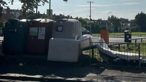 James Moore/LDRS A mattress and a bed frame dumped next to a row of large recycling bins in a public car park.