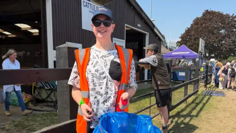 Liz Nice/BBC A woman in a baseball cap and orange hi-vis holds her blue litter bag