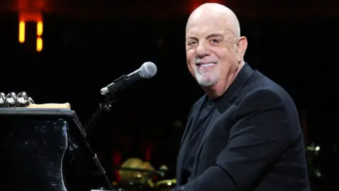 Billy Joel sitting at a piano and smiling at the camera