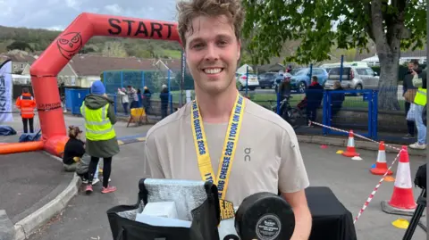 A man wearing a beige T-shirt is posing for a picture holding a wheel of cheese and a lunchbox. He is wearing a yellow medal and standing in front of a race finish line.