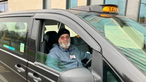 Waseem Saeed, a taxi driver who has worked in Stockport for 14 years, sat in his taxi looking disgruntled while parked at the station taxi rank. 