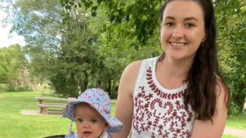 Handout A little baby girl with a colourful spotty hat on and blue eyes beside her mum who has her hand on her. Mum has brown long hair and brown eyes and is wearing a white round necked vest top with maroon on it. They are in a park with a bench and it is sunny.