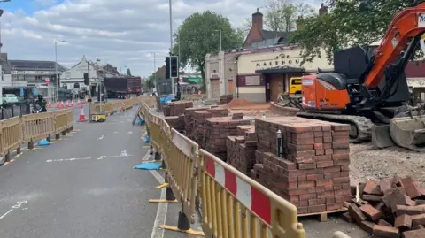 Piles of bricks can be seen behind plastic fencing on the right of the image, close to a small digger. Part of a road is fenced off on both sides due to construction work with cones marking the road further down the road.